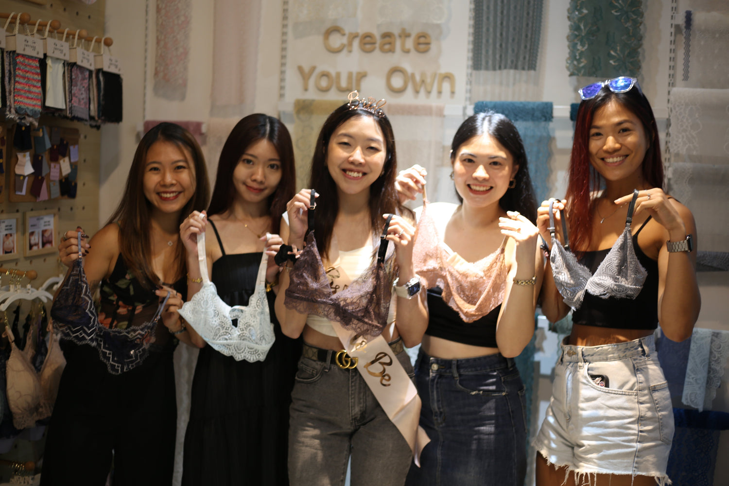 A smiling bride-to-be wearing a sash and tiara posing with four friends, holding their custom handmade lace bralettes during a Hens party sewing workshop.
