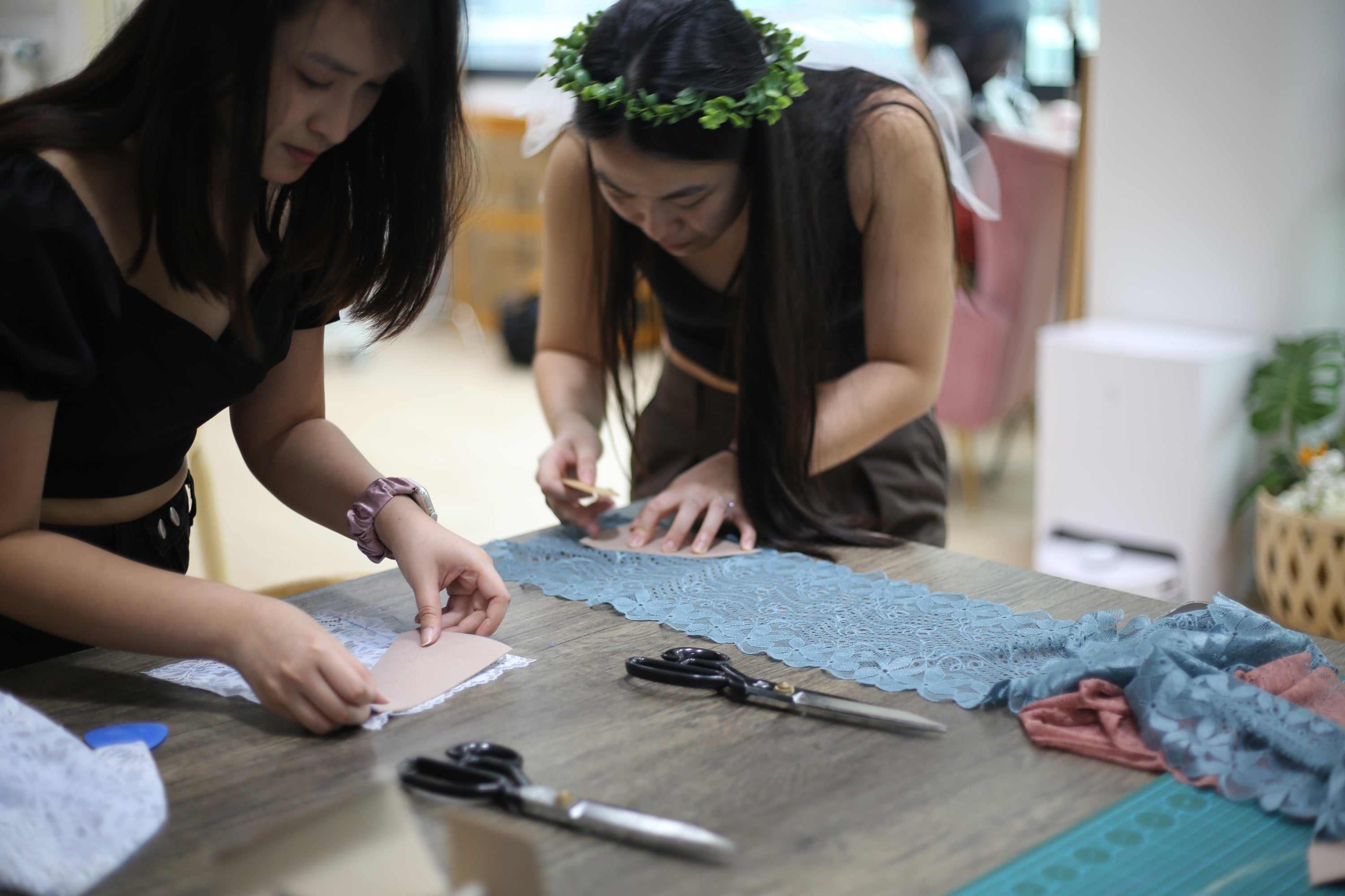 A bride-to-be wearing a leafy veil headband and a friend carefully cutting blue stretch lace on a cutting mat during a 5-pax Hens party bralette sewing workshop.