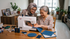 An elderly woman sits at a wooden table sewing with a white FEIYUE sewing machine, guided by a younger woman wearing a patterned hijab. Blue quilted fabric pieces are laid out on the table in a bright, modern home setting.