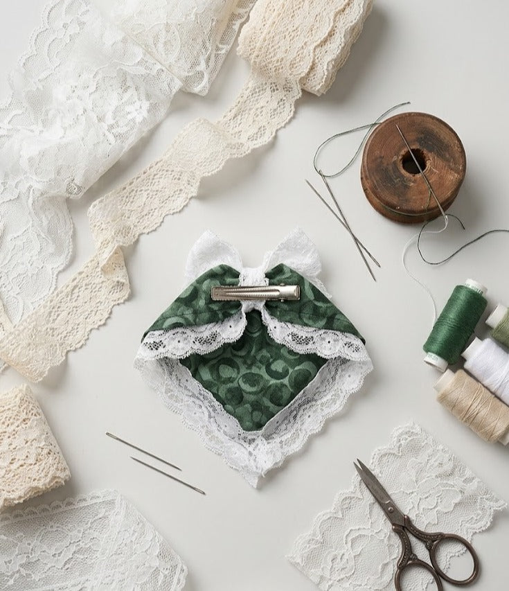 Flat lay of a DIY craft kit featuring a handmade green patterned fabric hair clip with a white lace bow and trim, surrounded by sewing needles, scissors, thread spools, and lace ribbons