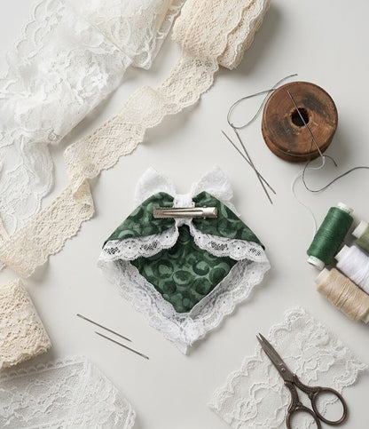 Flat lay of a DIY craft kit featuring a handmade green patterned fabric hair clip with a white lace bow and trim, surrounded by sewing needles, scissors, thread spools, and lace ribbons