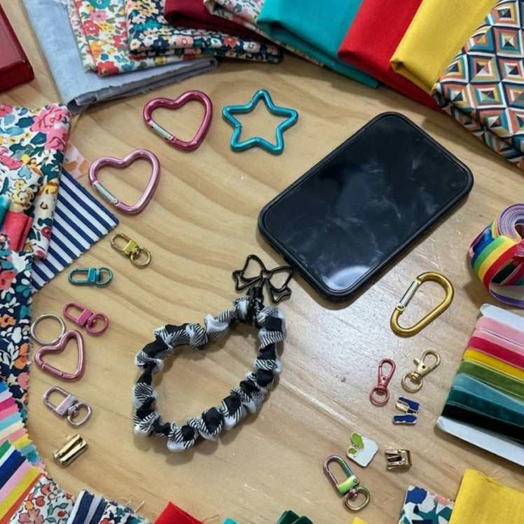 Flat lay of a DIY scrunchie phone wristlet sewing kit featuring a black and white checkered fabric wrist strap attached to a smartphone, surrounded by colorful fabric scraps, velvet ribbons, heart and star carabiners, and metal lobster clasps