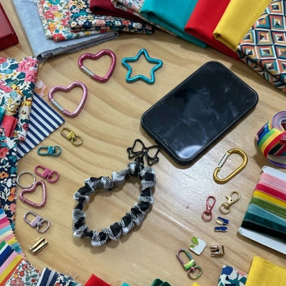 Flat lay of a DIY scrunchie phone wristlet sewing kit featuring a black and white checkered fabric wrist strap attached to a smartphone, surrounded by colorful fabric scraps, velvet ribbons, heart and star carabiners, and metal lobster clasps