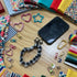 Flat lay of a DIY scrunchie phone wristlet sewing kit featuring a black and white checkered fabric wrist strap attached to a smartphone, surrounded by colorful fabric scraps, velvet ribbons, heart and star carabiners, and metal lobster clasps