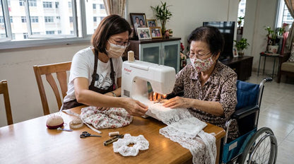 An elderly woman in a wheelchair uses a white sewing machine while a younger woman assists her at a wooden table in a home setting.
